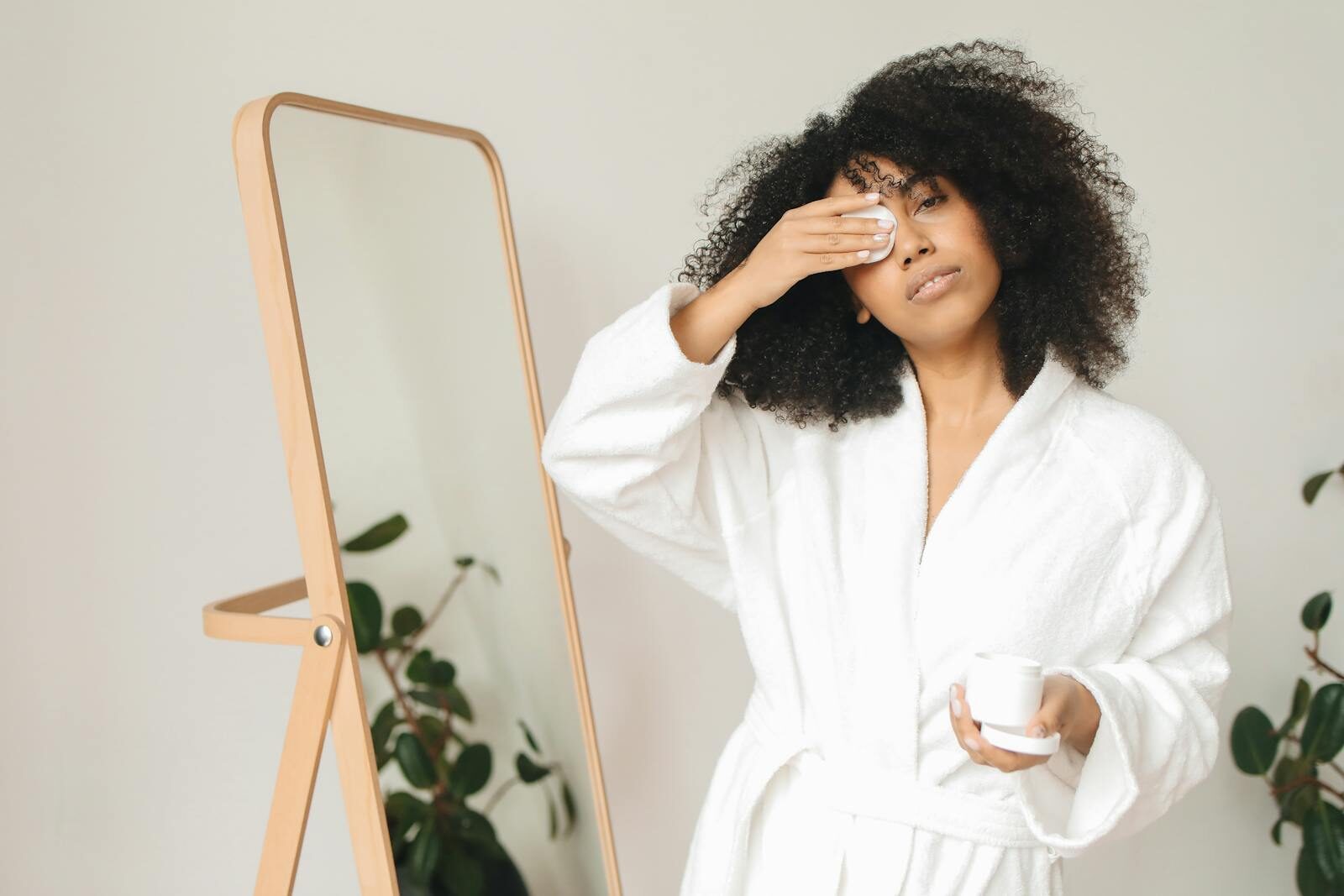 Woman in a white bathrobe using a cotton pad and holding a skincare container indoors.